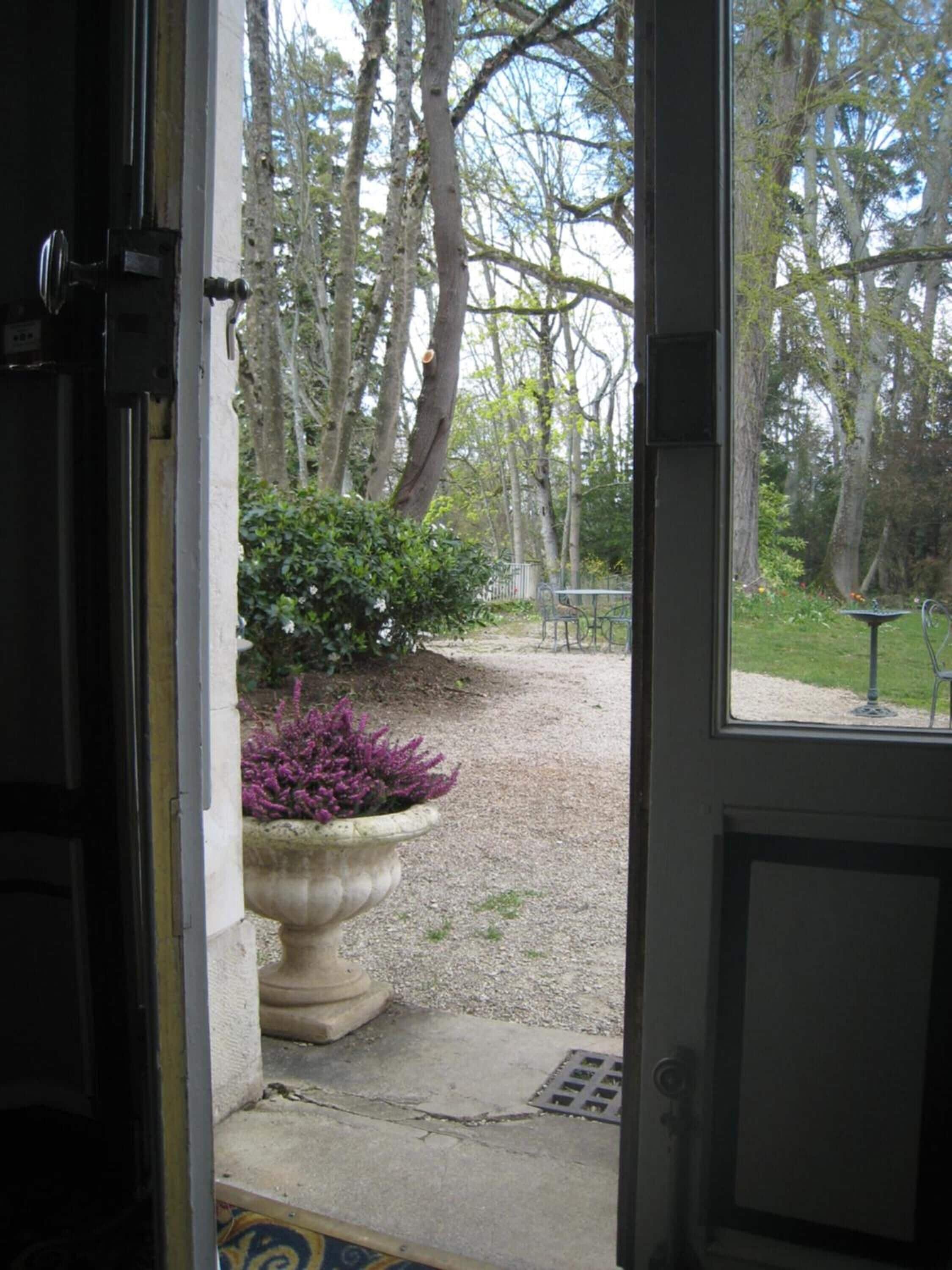 An open door leading to a gravel path with a large planter of purple flowers, surrounded by trees.