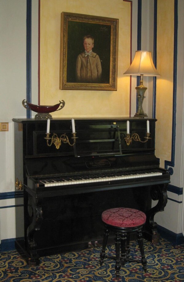 A piano with a stool in front, a framed portrait above, and a lamp to the right.