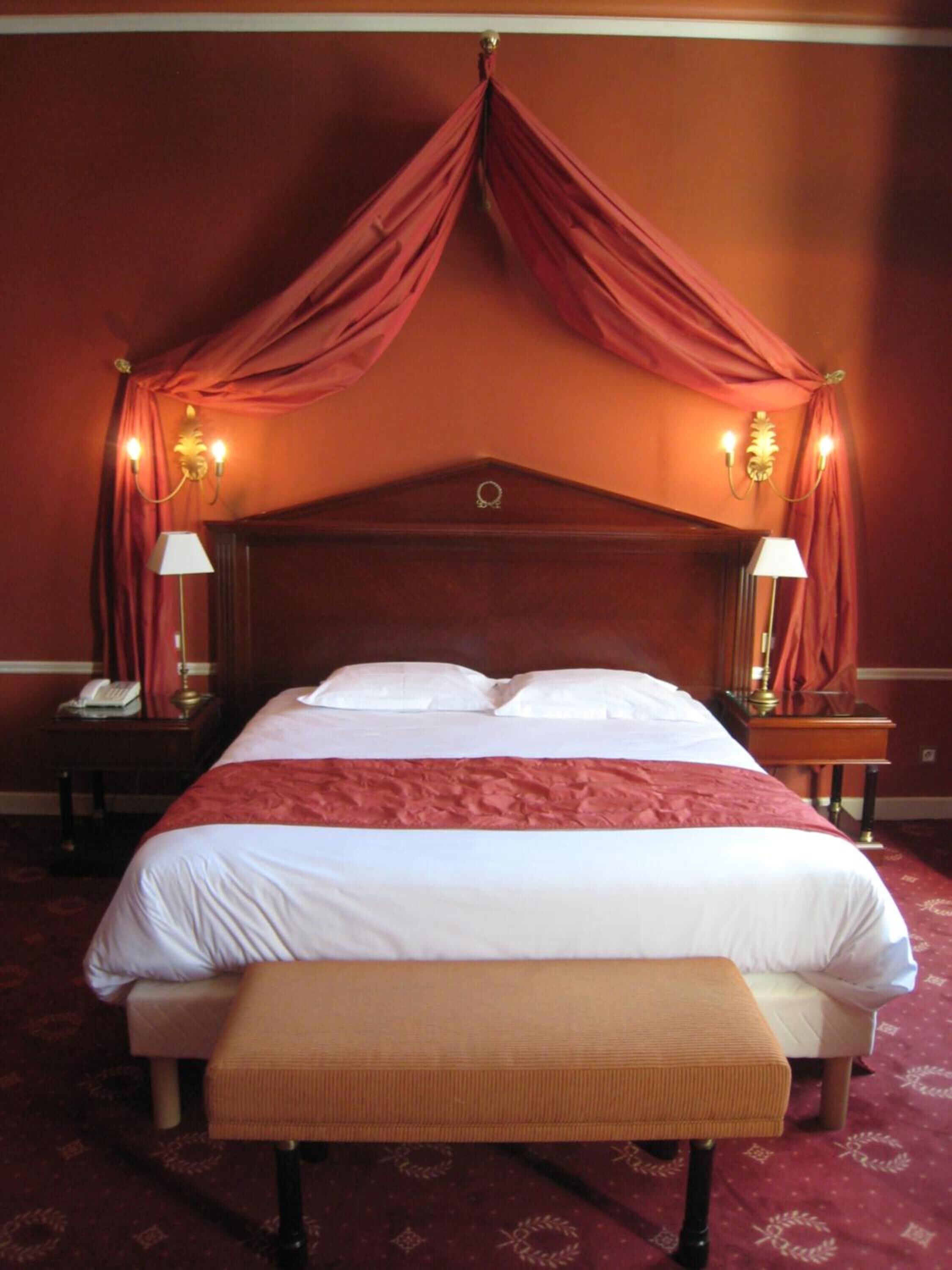 Habitación de hotel con una cama grande, un banco, dos mesitas de noche con lámparas y una cama con dosel y cortinas.