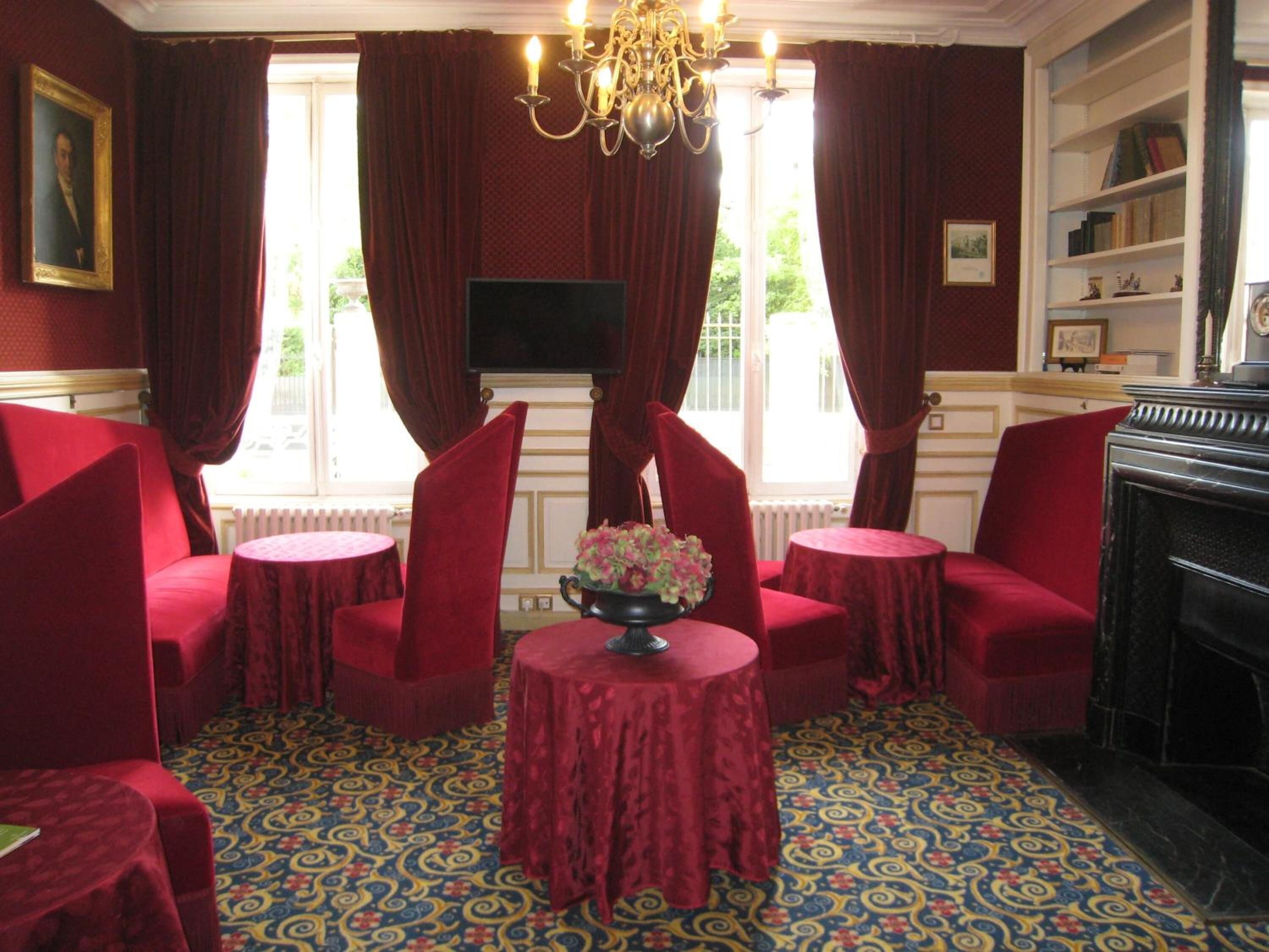 A room with red upholstered chairs, a round table with a vase, patterned carpet, a fireplace, and a bookshelf.