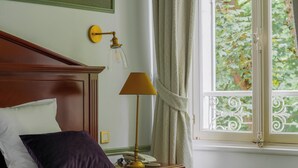 Un dormitorio con cabecera de madera, cama con almohadas blancas y púrpuras, mesita de noche con lámpara y ventana con vista a zonas verdes.
