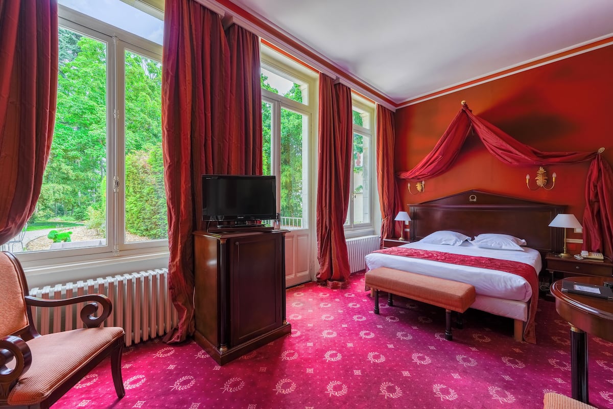 Een hotelkamer met een groot bed, een televisie, een bankje en uitzicht op groen door de ramen.