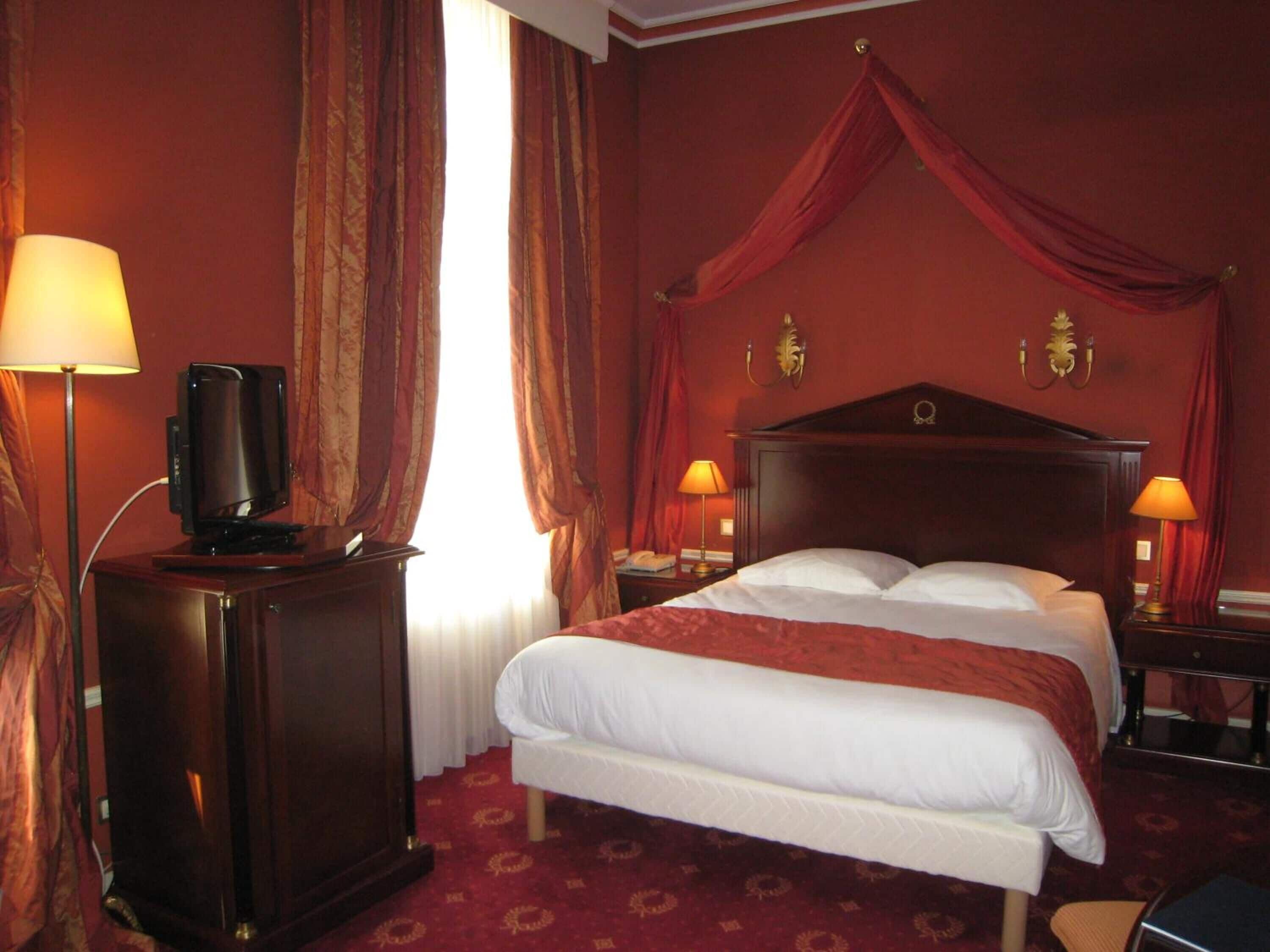 A hotel room with a large bed, two bedside lamps, a TV on a wooden cabinet, and a window with curtains.