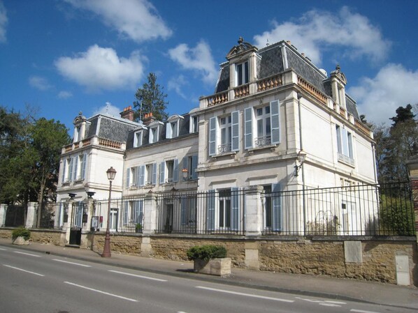 Un edificio grande e histórico con fachada simétrica, múltiples ventanas con contraventanas y una entrada destacada con pórtico.