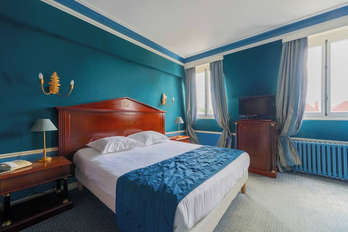 Een hotelkamer met een groot bed, een houten nachtkastje, een bureau met een lamp, een televisie en een raam met gordijnen.