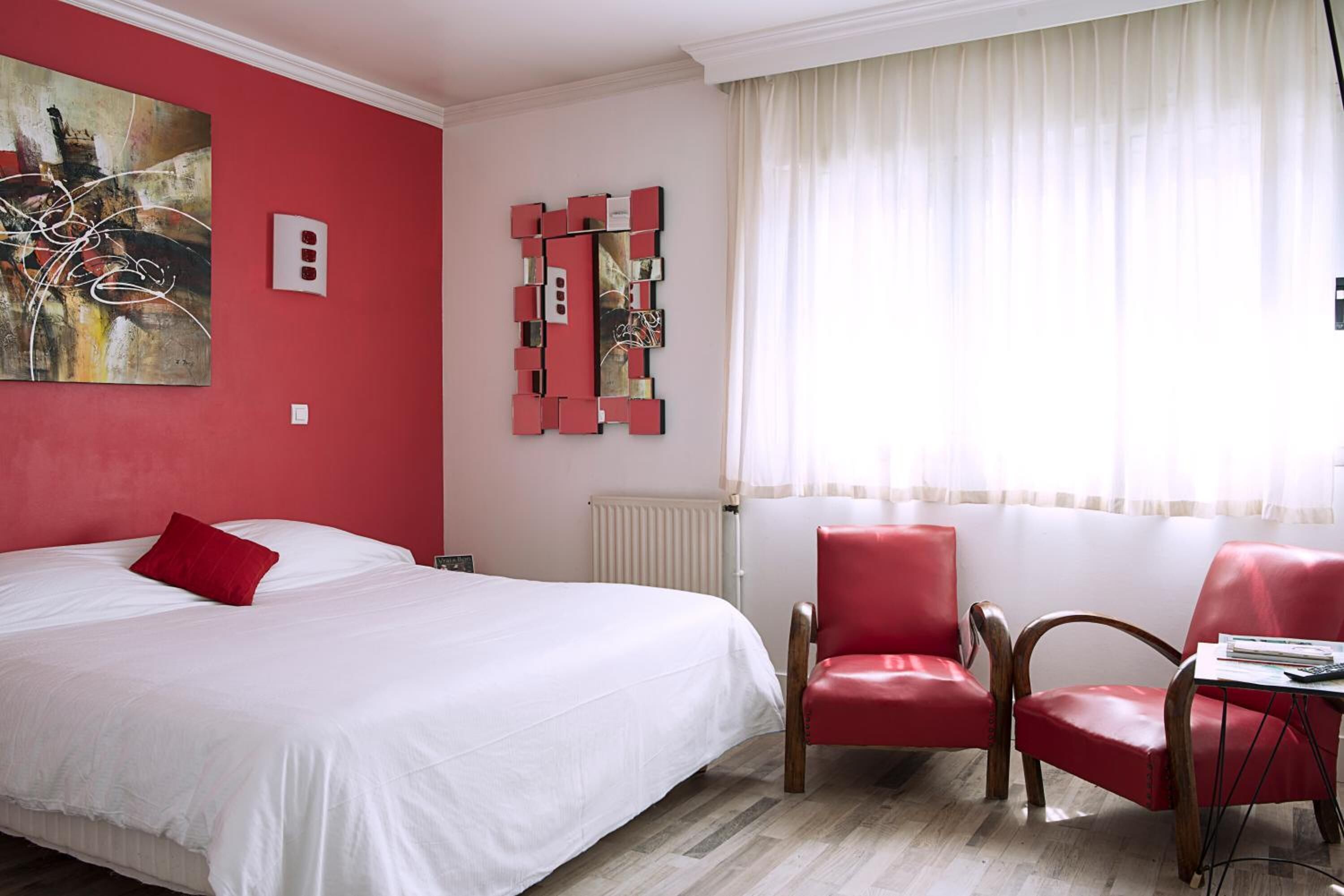 A hotel room with a large bed, red chairs, a table, a red abstract painting, and a red geometric wall art piece.