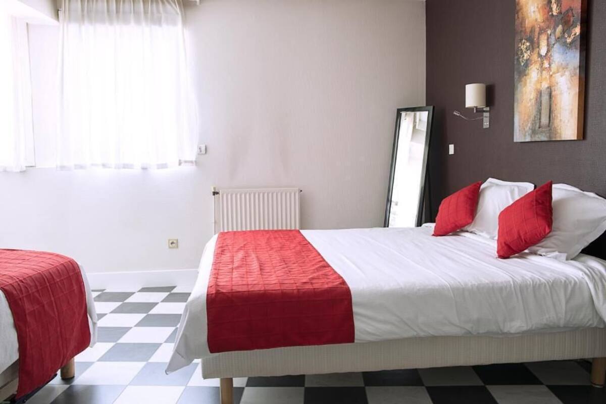 Ein Hotelzimmer mit Schachbrettboden, einem Bett mit weißer und roter Bettwäsche, einem Heizkörper, einem Bild an der Wand und einem Fenster mit Vorhängen.