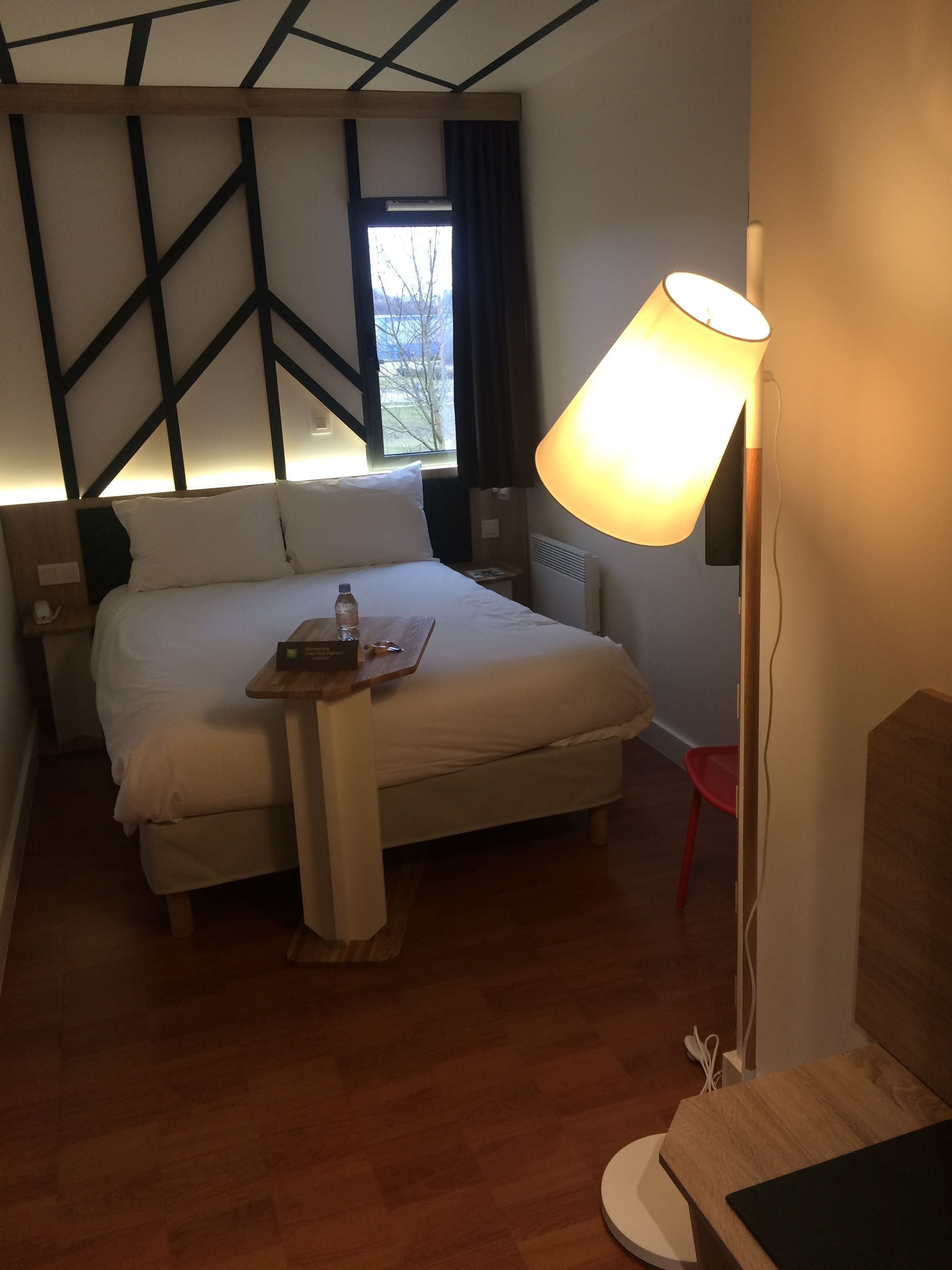 A hotel room with a bed, bedside table, lamp, and a view of the outdoors.