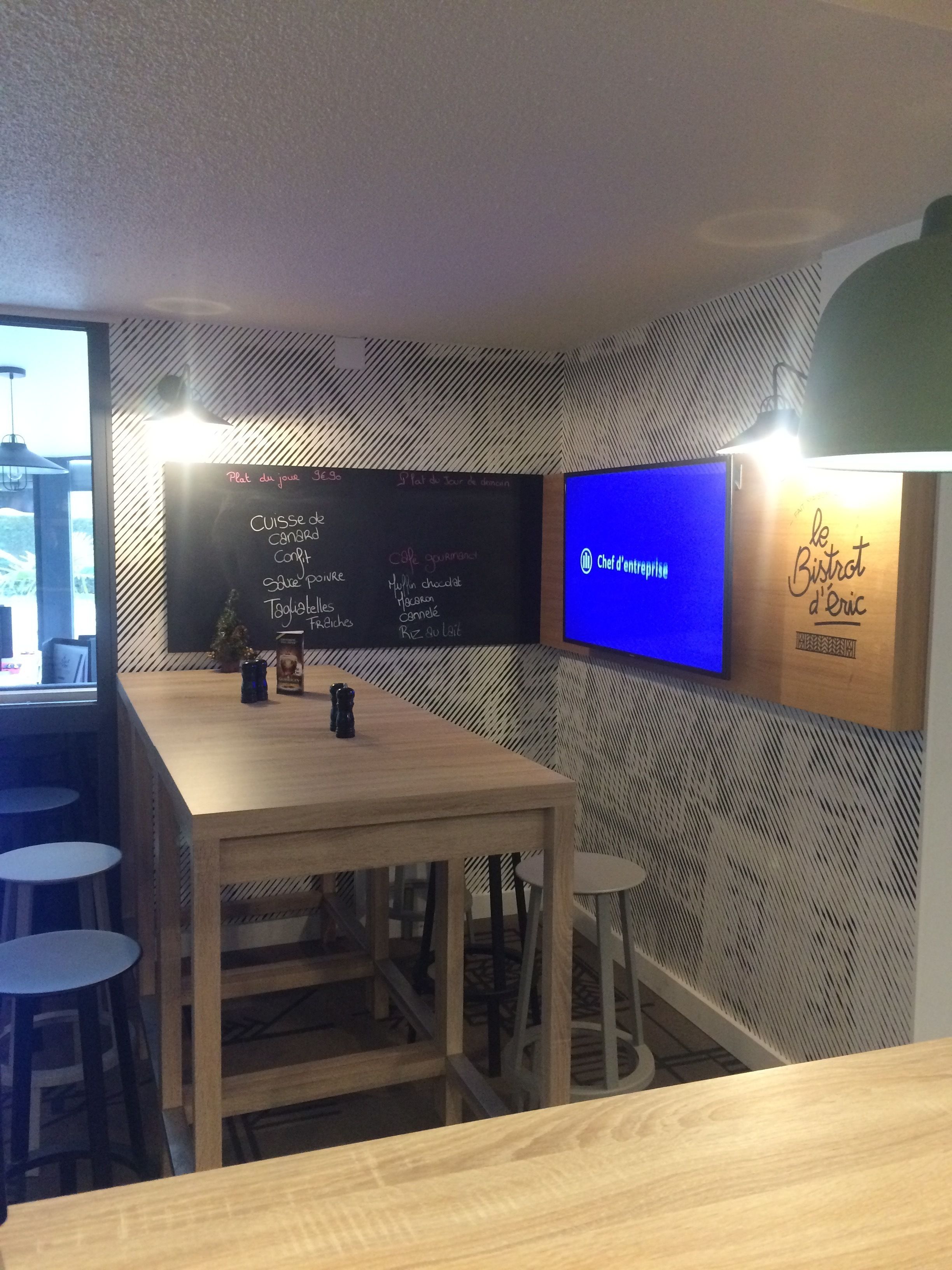 L’intérieur d’un restaurant avec une table en bois, des tabourets, un menu écrit à la craie et un écran de télévision.