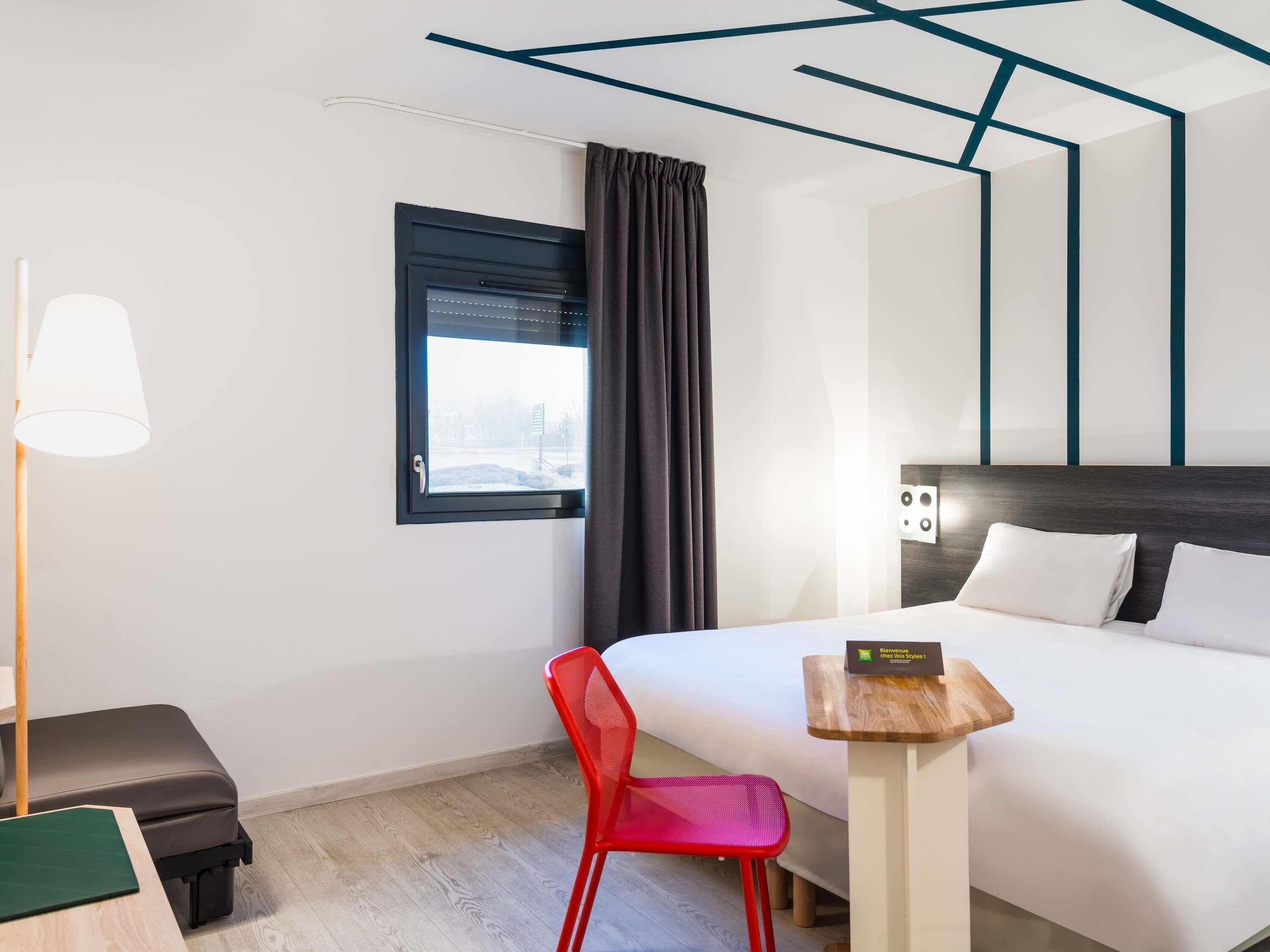 Une chambre d’hôtel avec un lit, un bureau, une chaise, une lampe et une fenêtre avec des rideaux.