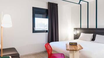 Une chambre d’hôtel avec un lit, un bureau, une chaise, une lampe et une fenêtre avec des rideaux.