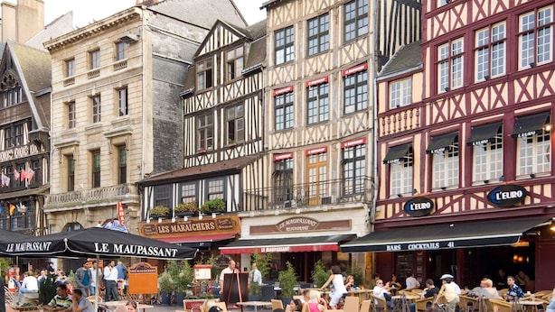 A European street with outdoor seating at cafes, historic buildings, and signs for 'LE MAUPASSANT' and 'L'EURO'.