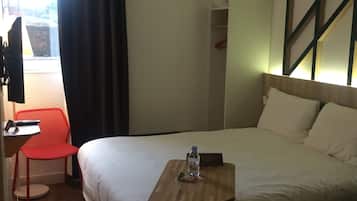 Et hotelværelse med en seng, et skrivebord med stol, et lille bord med en flaske og et vindue med gardiner.