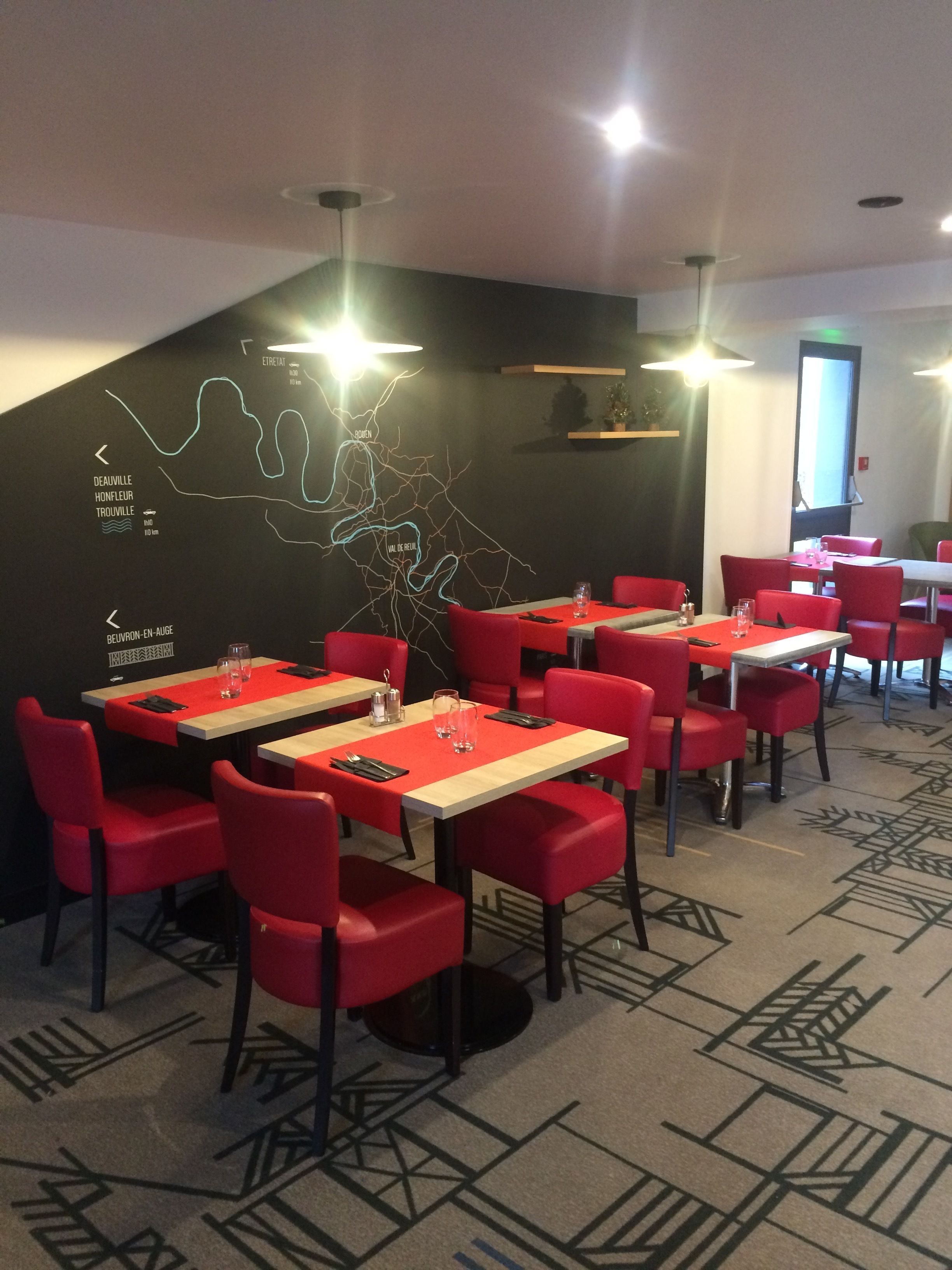 Een restaurant met rode stoelen en tafels, een zwarte muur met een kaart, en verlichting aan het plafond.