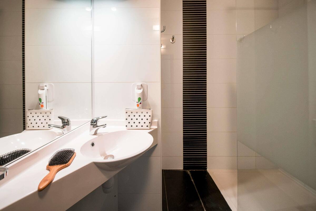A modern bathroom with a white sink, two mirrors, a wooden-handled brush, and a black shower drain.