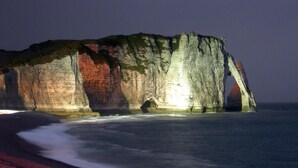A large rock formation with a natural arch, located near a beach with red sand.