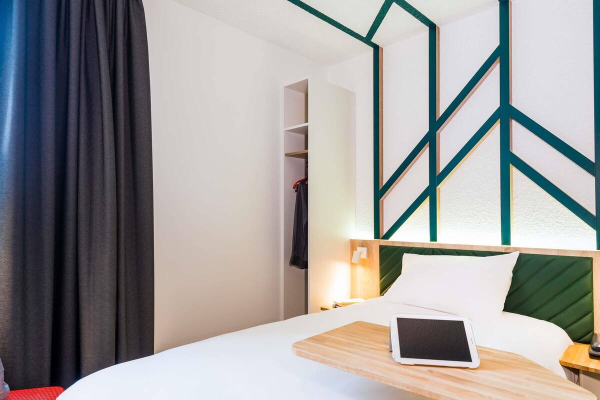 A hotel room with a bed, a bedside table with a tablet, a red chair, and a geometric wall design.
