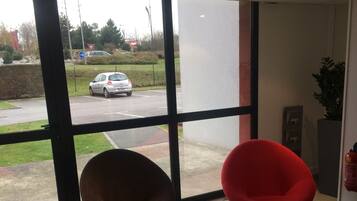 A waiting area with two red chairs, a brown chair, and a small table with magazines. Large windows provide a view of a parking lot and a grassy area outside.