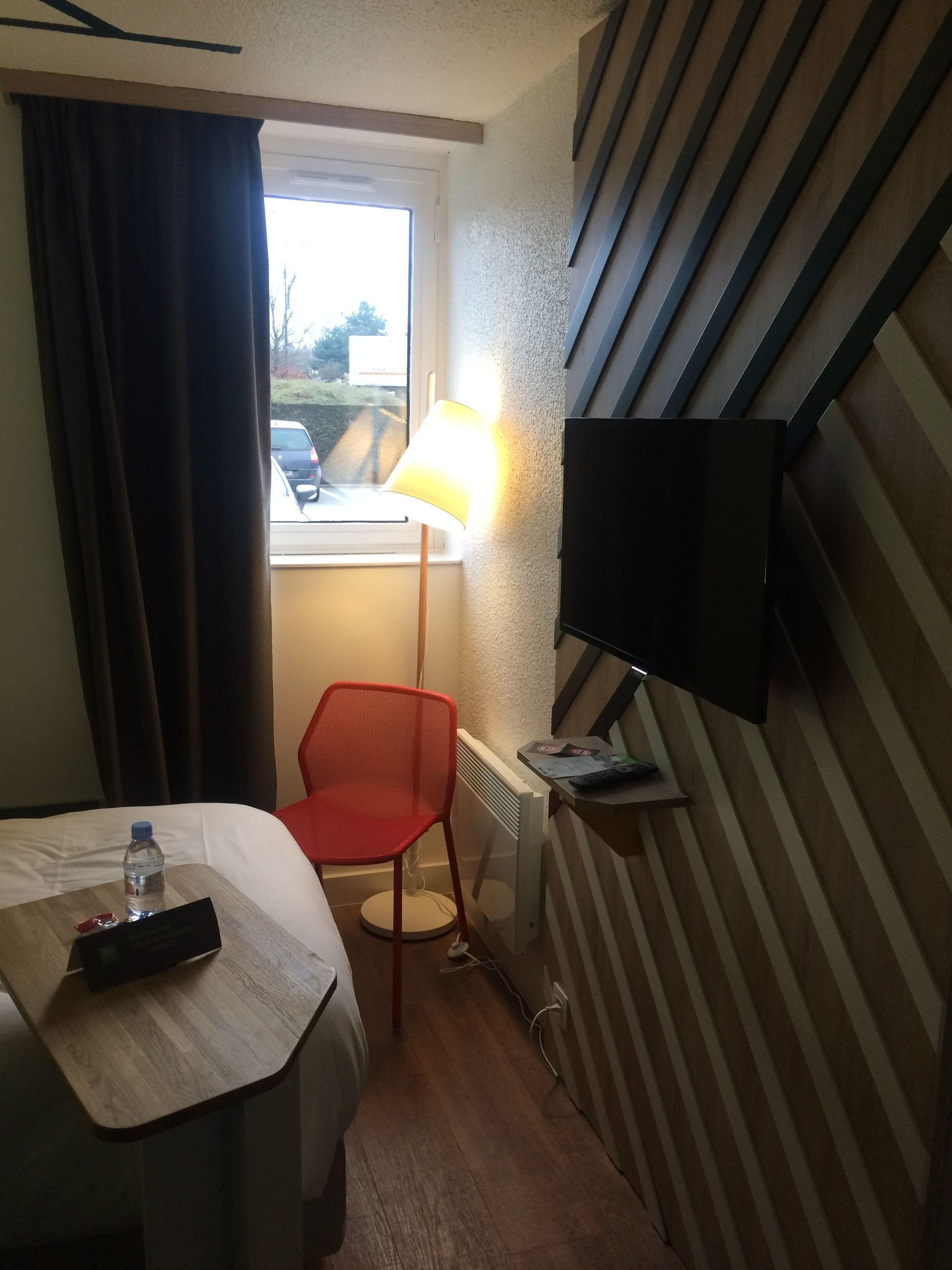 Habitación de hotel con cama, una mesita con un control remoto, una silla roja, una lámpara y un televisor montado en la pared.
