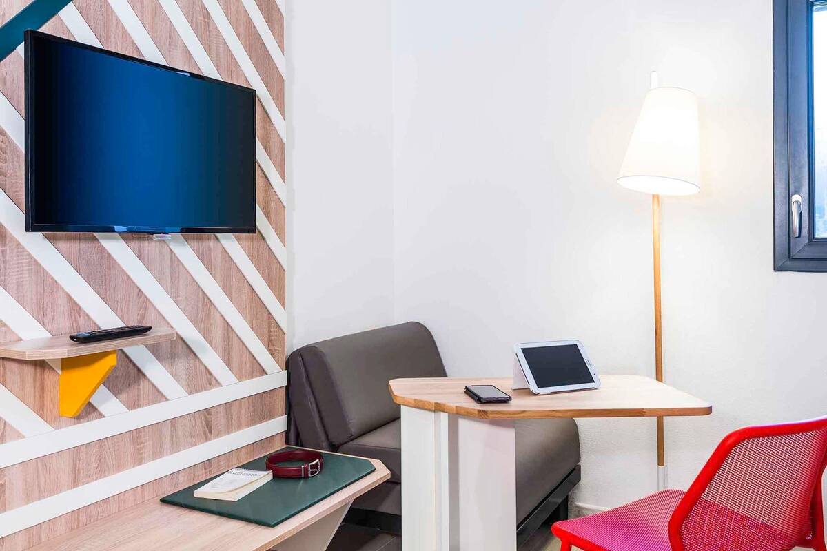 A modern hotel room with a flat-screen TV mounted on a wooden wall, a desk with a lamp, a chair, and a red chair.