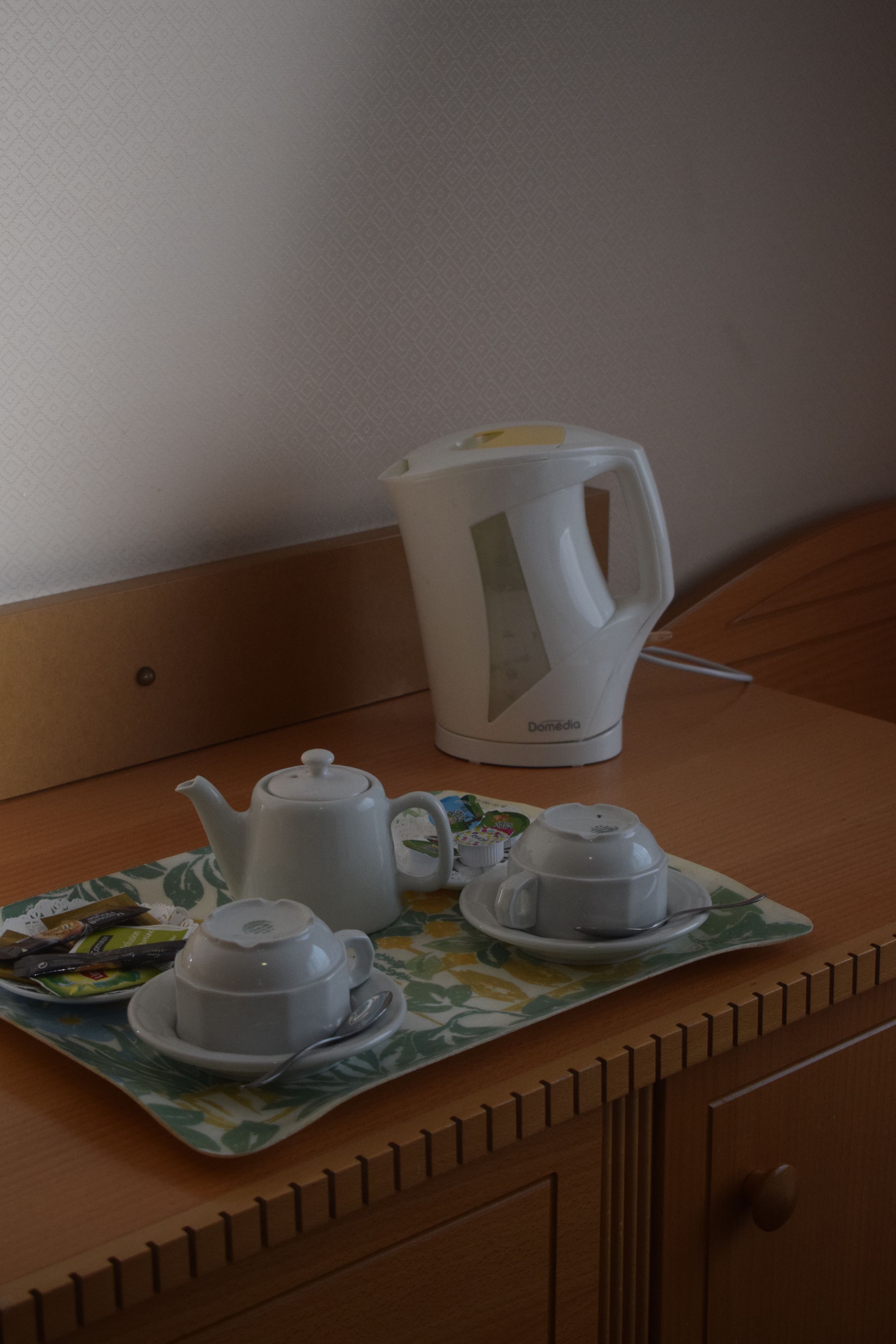 A wooden table with a white teapot, a white coffee maker, and two white cups on a floral placemat.