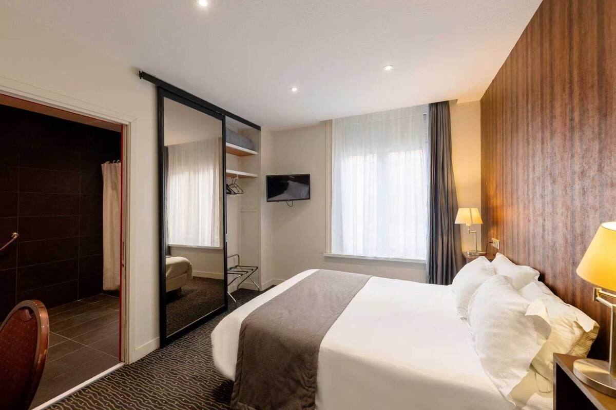 Ett hotellrum med en säng, en TV, ett skrivbord med en lampa, en dusch och en garderob.