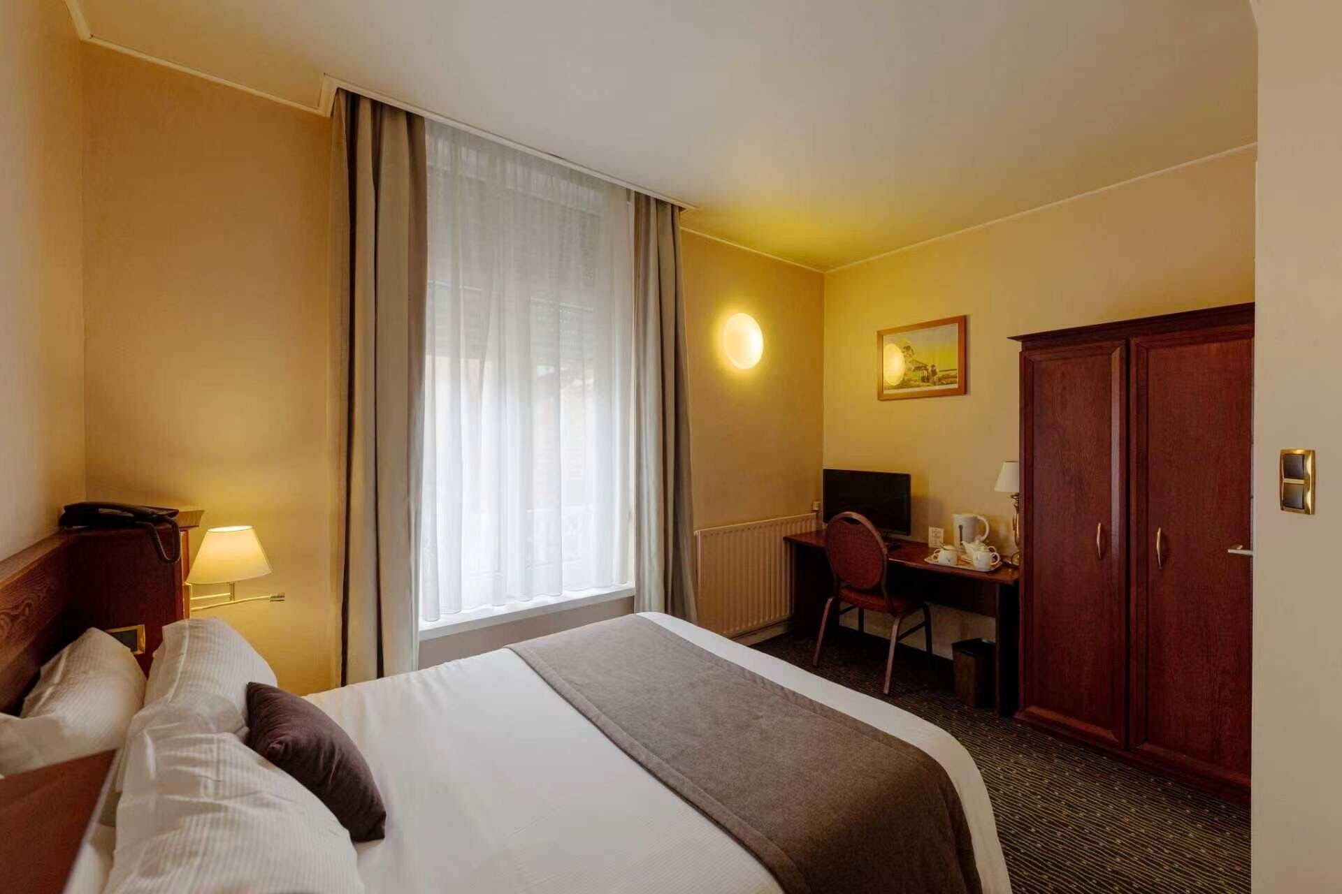 Une chambre d’hôtel avec un lit, un bureau, une chaise, une armoire et une fenêtre avec des rideaux.