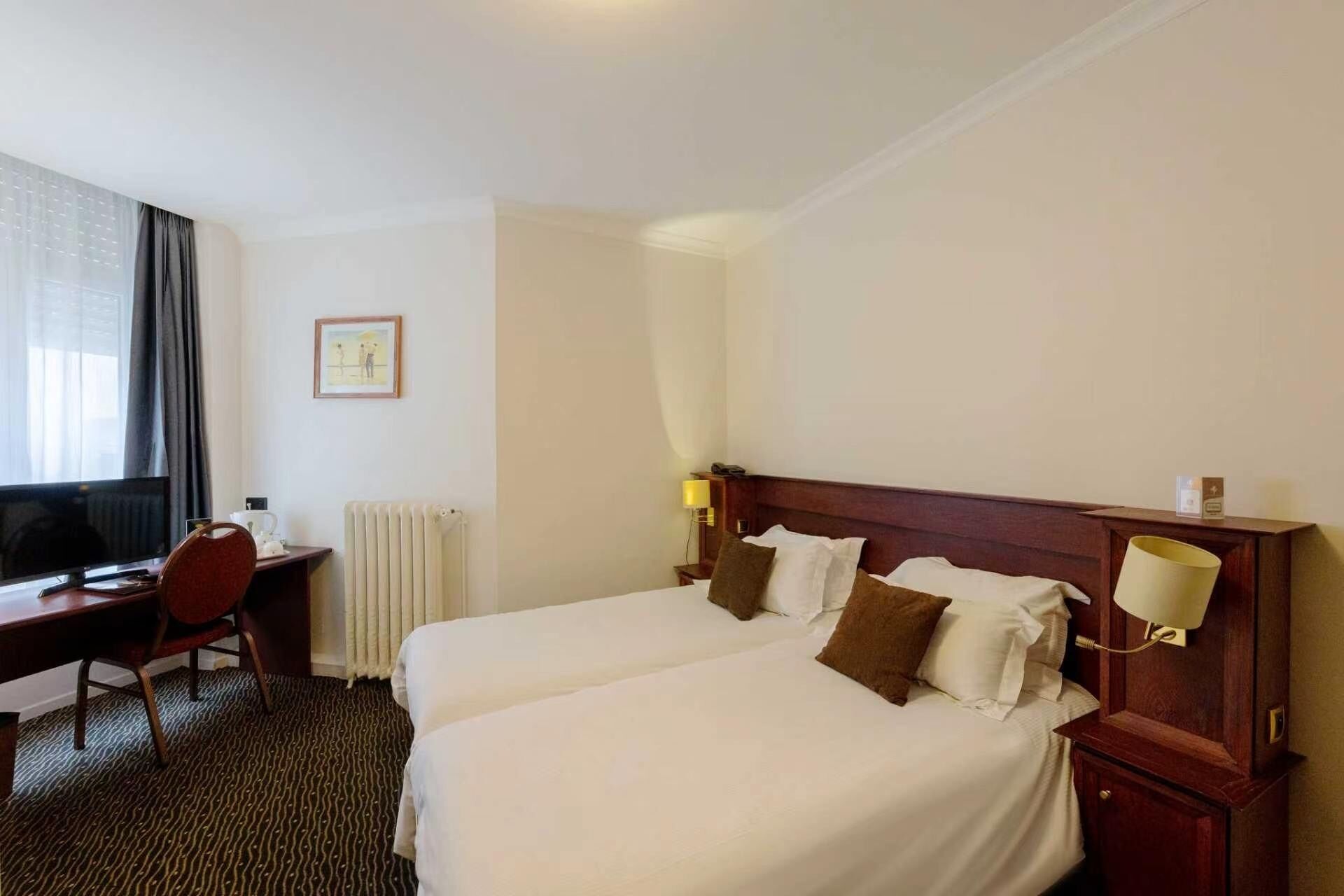 Habitación de hotel con dos camas, un escritorio, una silla, un radiador, un cuadro en la pared y una ventana con cortinas.