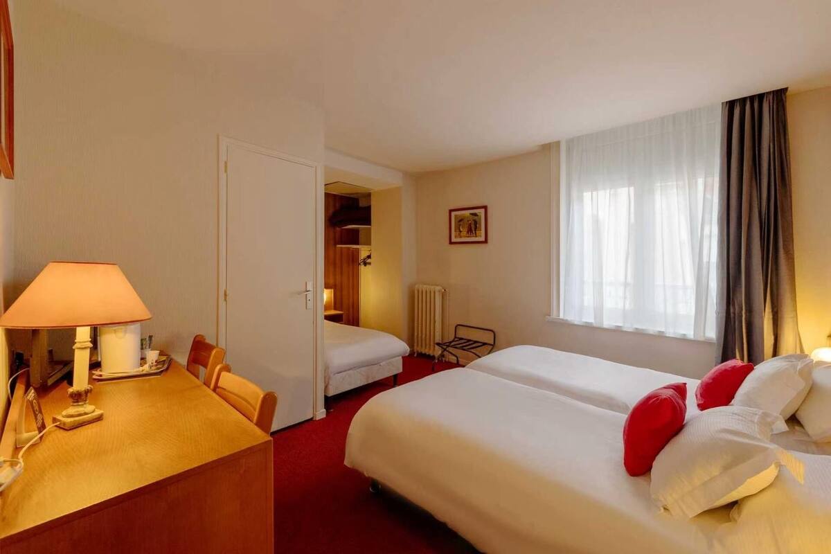 Ett hotellrum med två sängar, ett skrivbord med en lampa, en stol, ett element och ett fönster med gardiner.