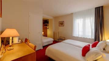 Habitación de hotel con dos camas, un escritorio con lámpara, una silla, un radiador y una ventana con cortinas.