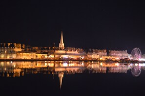 Ein beleuchtetes Stadtbild bei Nacht, mit einem markanten Turm und einem Riesenrad, dessen Spiegelung sich im Wasser zeigt.