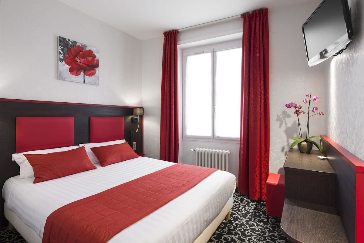 A hotel room with a bed, red curtains, a TV, a plant, and a radiator.