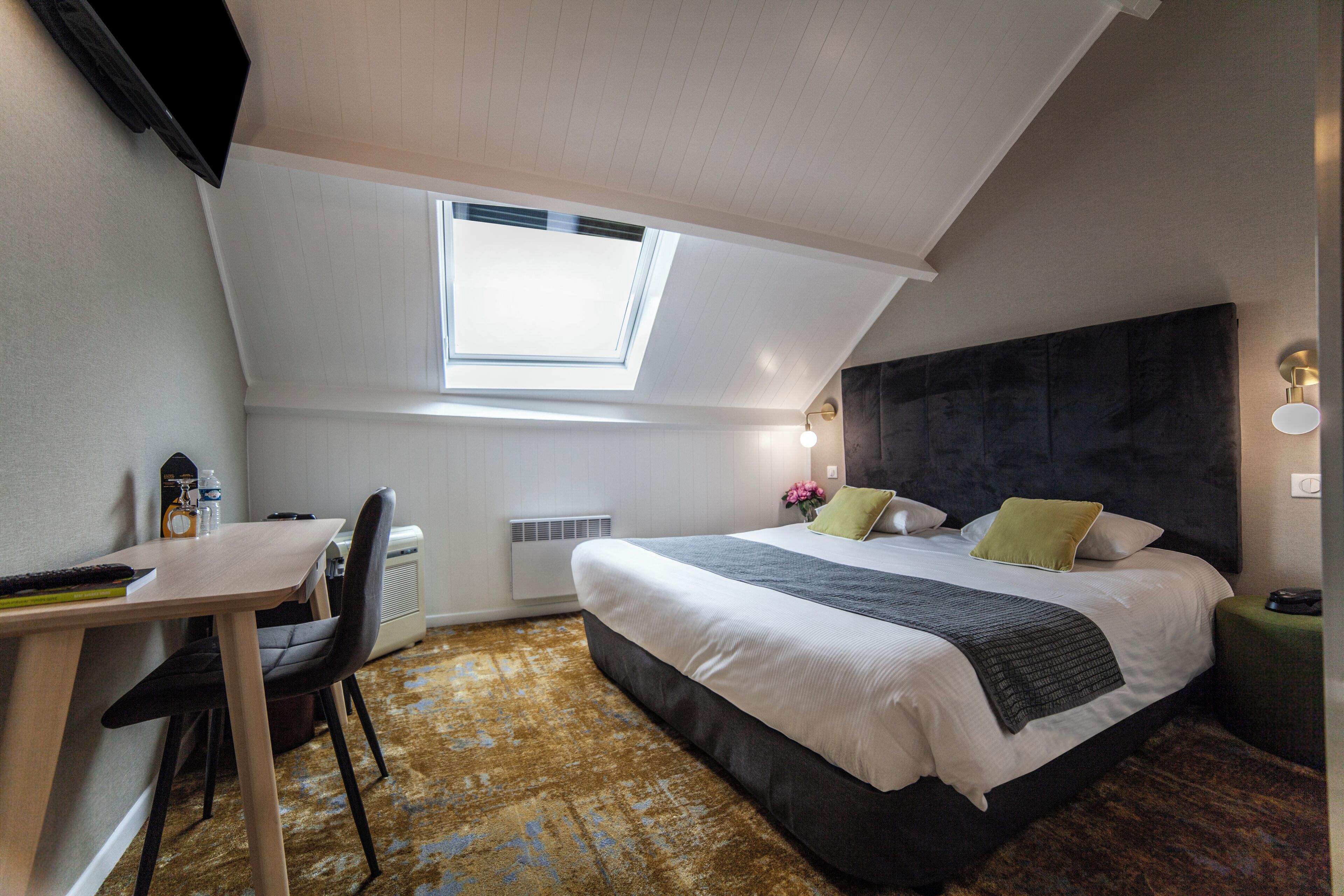 A bedroom with a skylight, a bed with pillows, a desk with a chair, and a radiator.