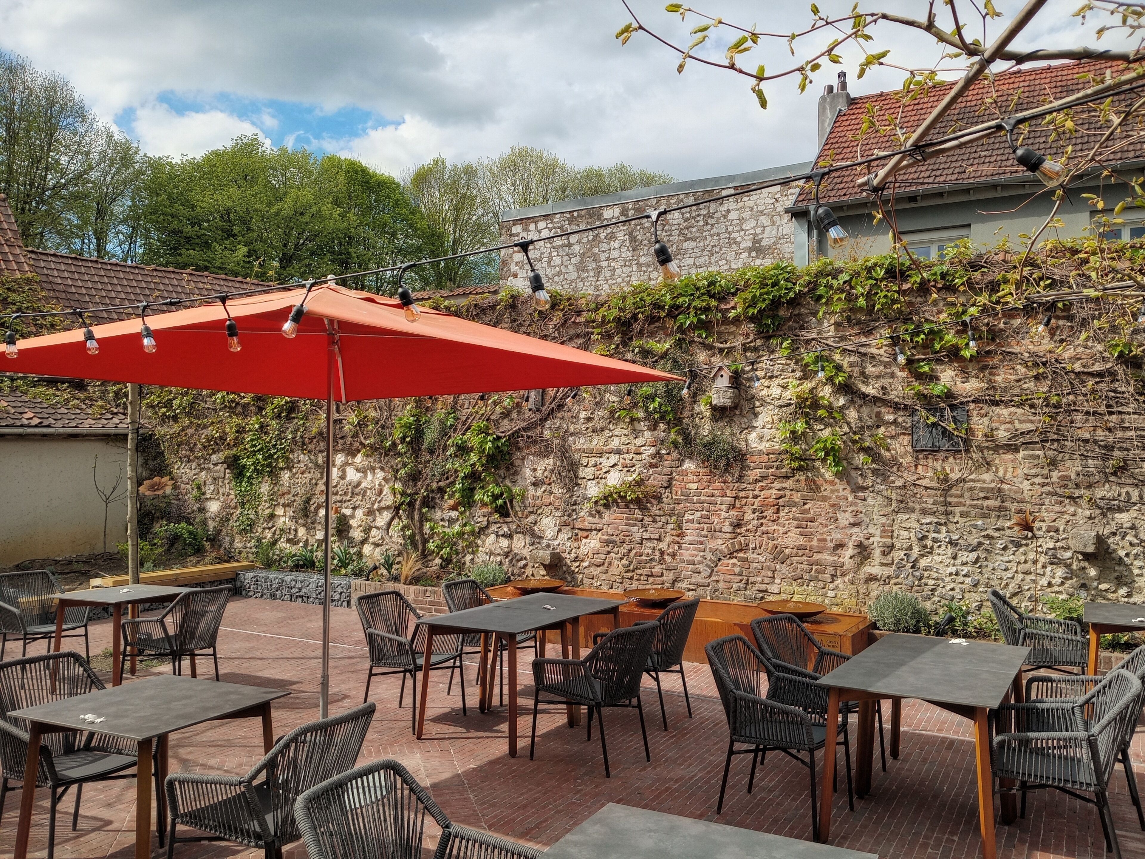 Une terrasse extérieure avec un sol en briques, des chaises et des tables en osier, le tout installé sous un parasol rouge.