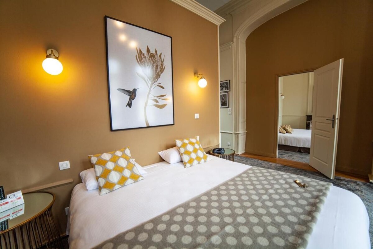 A hotel room with a large bed, two wall sconces, a framed artwork, and a door leading to another room.