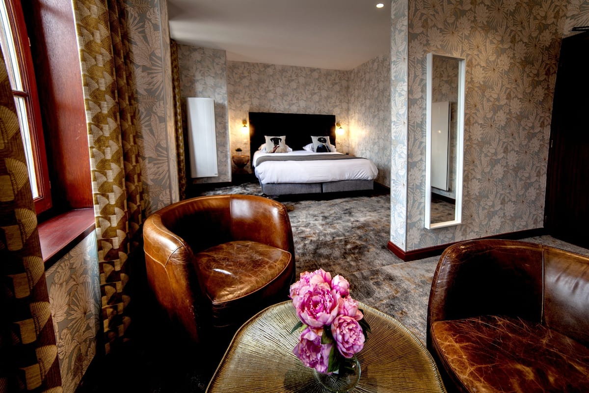 A hotel room with a large bed, two bedside lamps, a mirror, a leather armchair, a small round table with a vase of flowers, and patterned wallpaper.