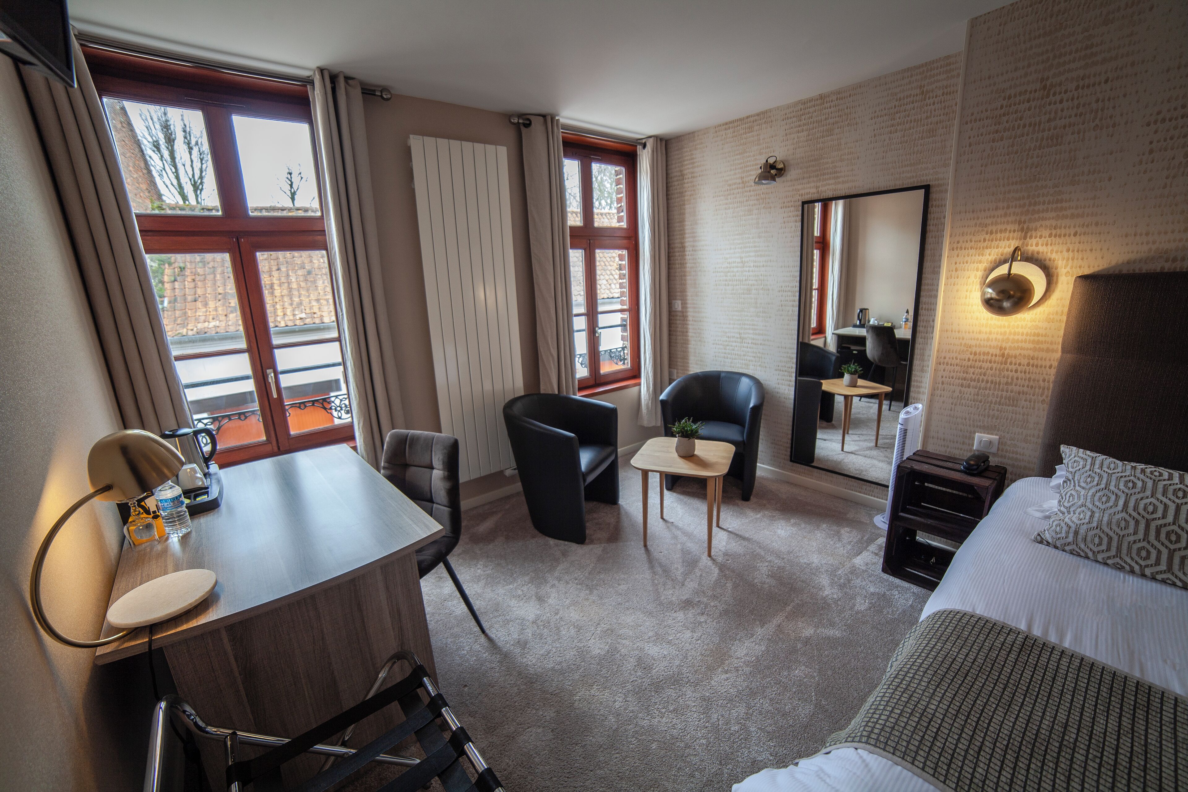 Et hotelværelse med skrivebord, to stole, en seng, et lille bord med en plante og et stort spejl.