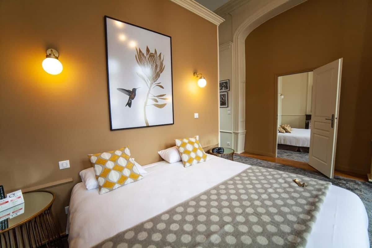 A hotel room with a large bed, two wall sconces, a framed artwork, and a door leading to another room.