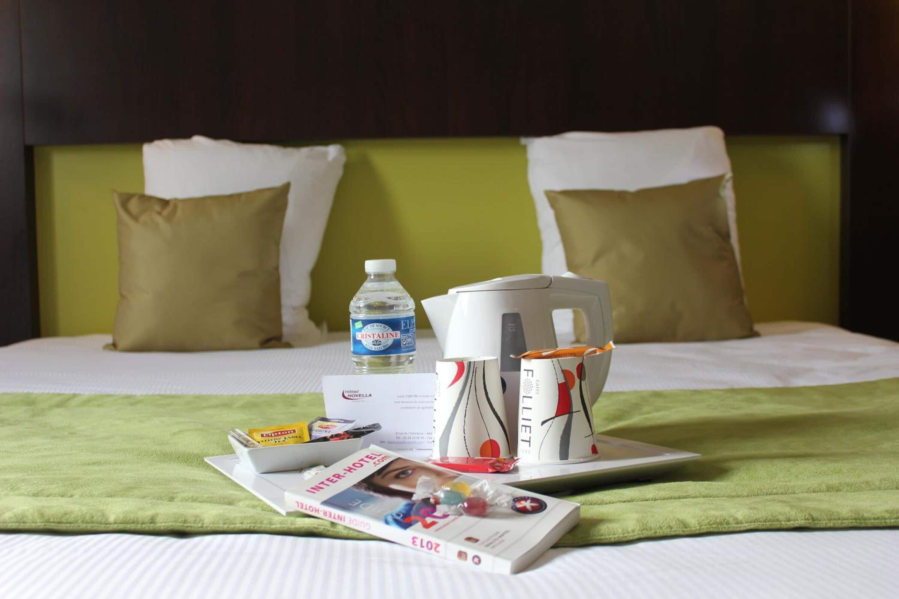 A neatly made bed with a green blanket, a bottle of water, a coffee maker, and a magazine on a tray.