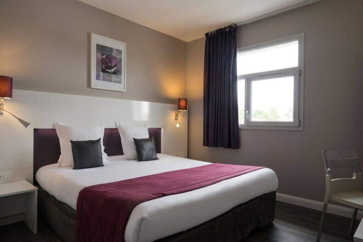 Een hotelkamer met een groot bed, twee bedlampen, een raam met gordijnen en een lijst met een afbeelding aan de muur.