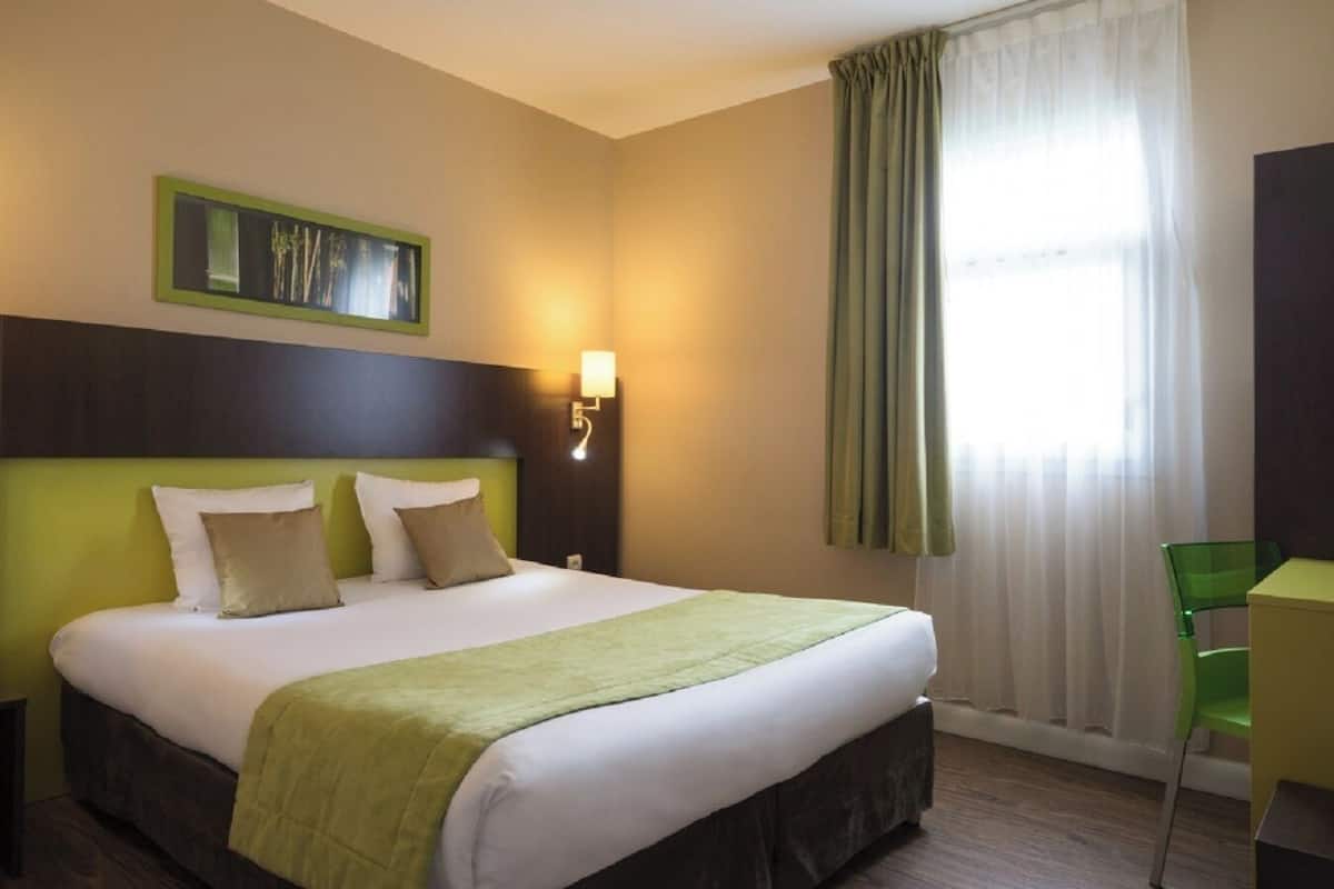 Hotelkamer met een bed, bureau, stoel, nachtkastje, wandlamp en raam met gordijnen.