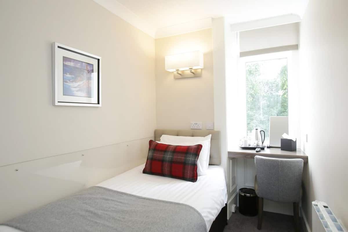 A hotel room with a bed, a desk, a chair, a window, and a wall-mounted light.