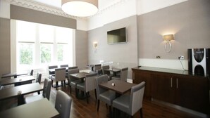 A modern dining area with tables and chairs, a large window, and a television mounted on the wall. - Ambassador Hotel (Glasgow)