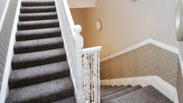 Una escalera con alfombra gris, barandilla blanca y una pared de color claro con un aplique de luz redondo.