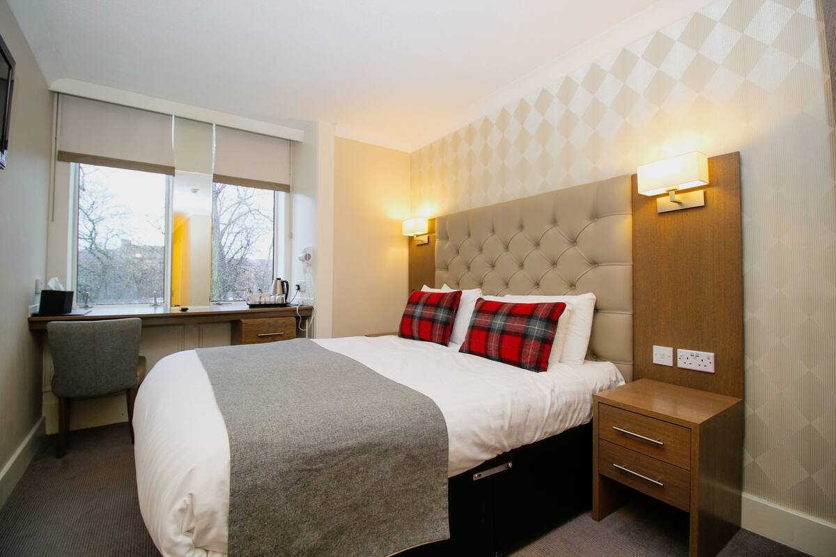A hotel room with a large bed, a desk, and a chair. There are two wall-mounted lamps on the bed's headboard.