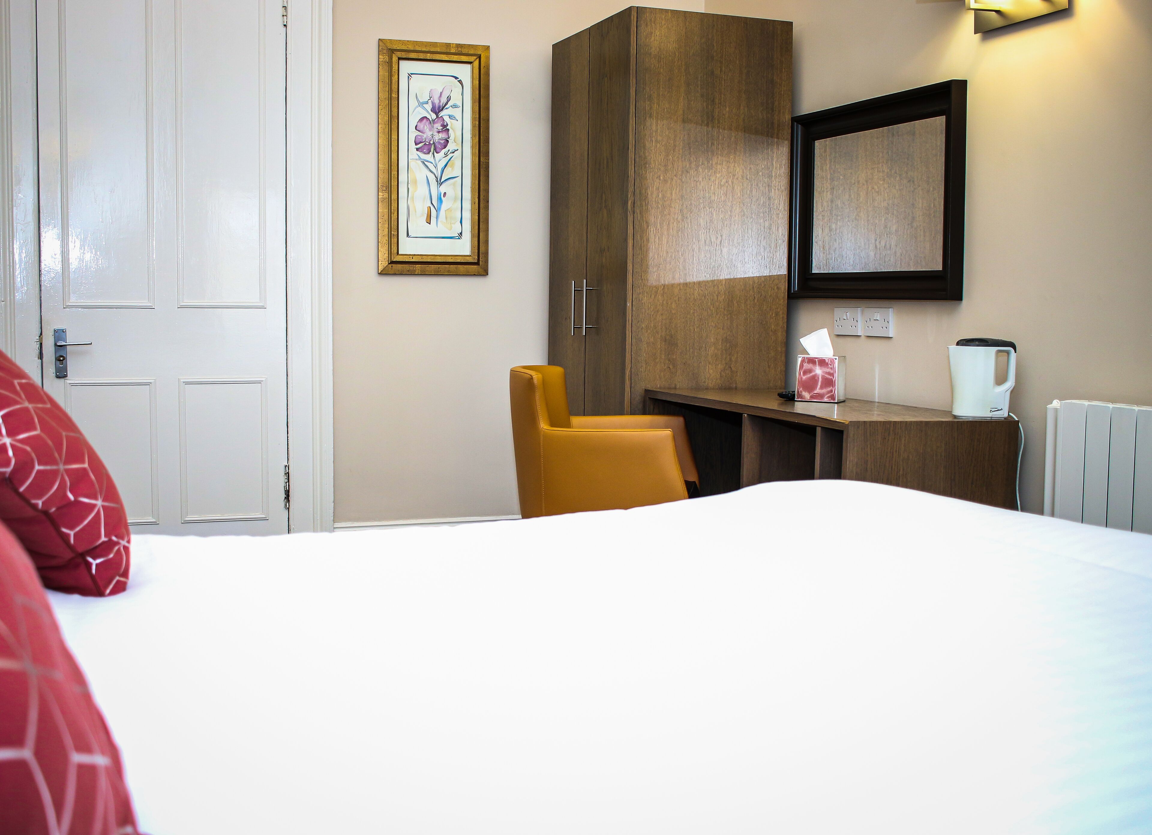 Una habitación de hotel con una cama, una silla amarilla, un armario de madera, una obra de arte floral enmarco, una mesita de noche con una caja de pañuelos y una tetera, y una puerta blanca.