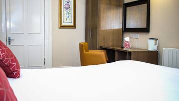Una habitación de hotel con una cama, una silla amarilla, un armario de madera, una obra de arte floral enmarco, una mesita de noche con una caja de pañuelos y una tetera, y una puerta blanca.