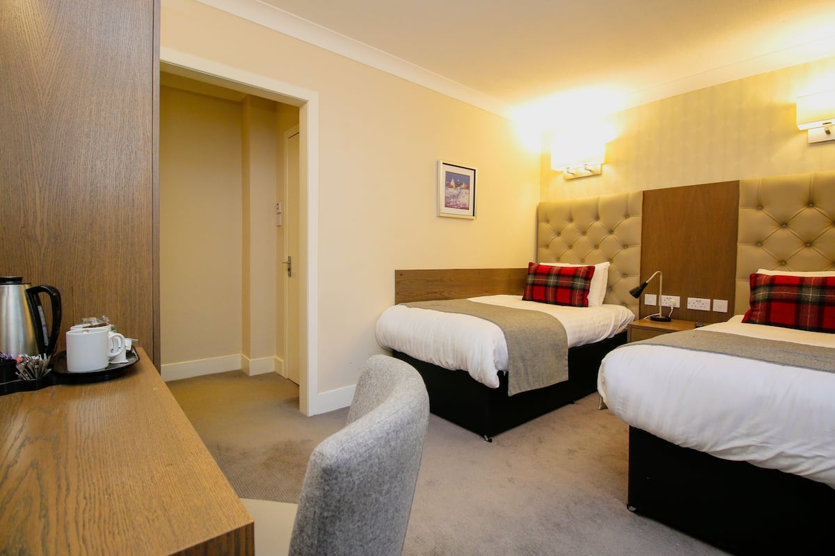 A hotel room with two beds, a wooden desk with a kettle, and a framed picture on the wall.