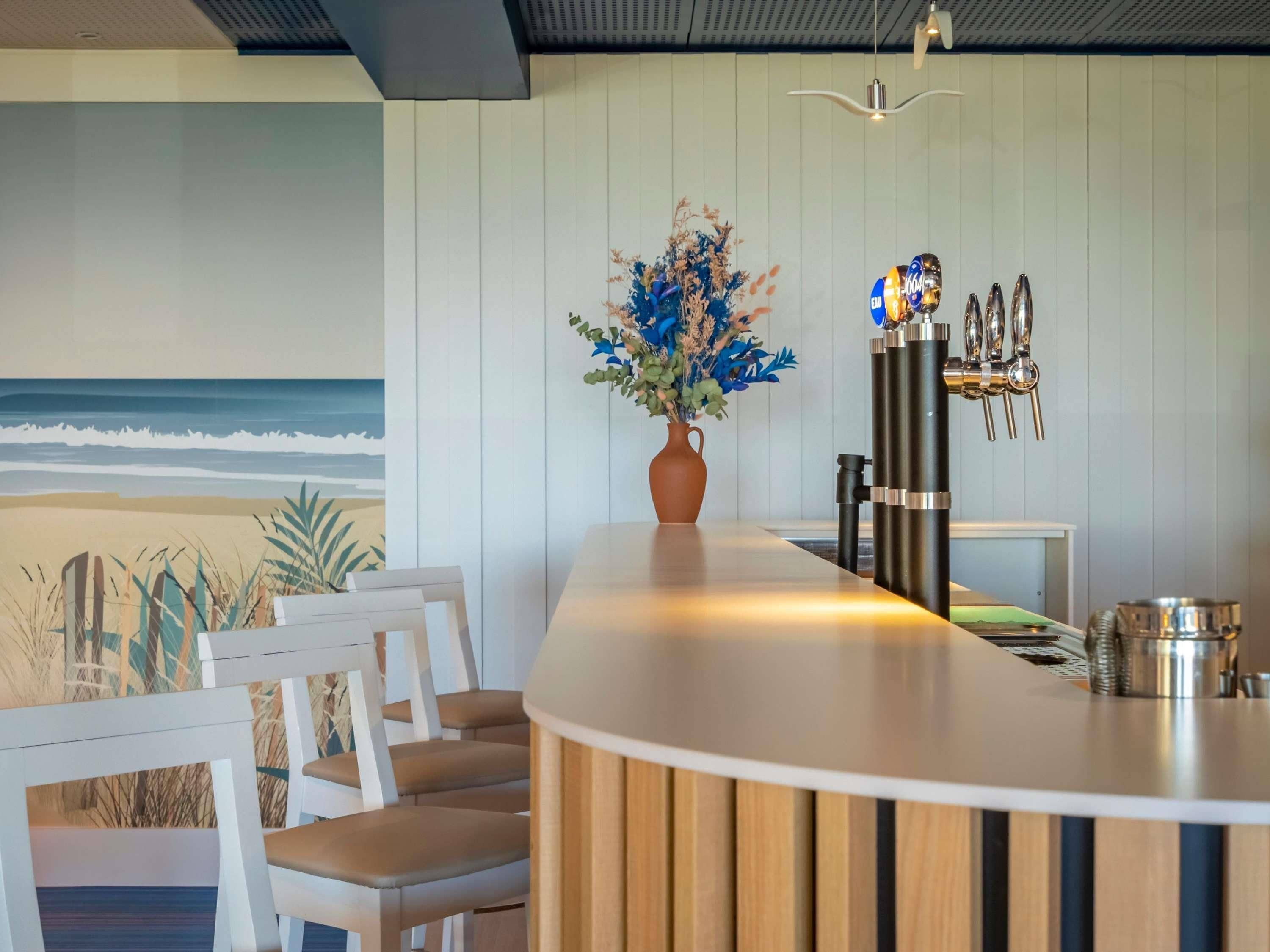 A modern kitchen with a wooden countertop, white chairs, a vase with flowers, and a view of the ocean.