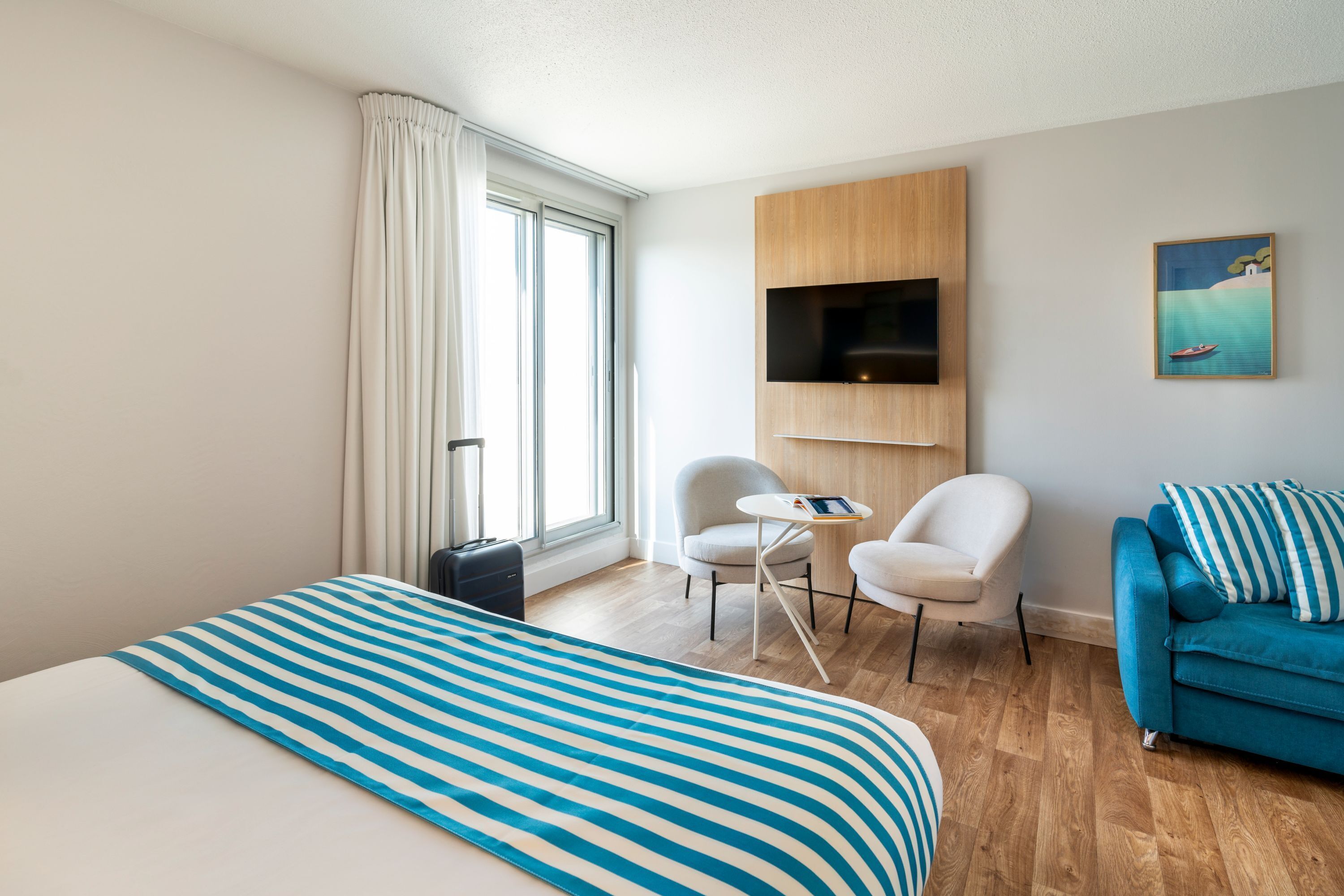 Une chambre d’hôtel avec un grand lit, une couvre-lit à rayures bleues, un canapé bleu, un téléviseur fixé au mur et un tableau représentant un bateau sur la mer.